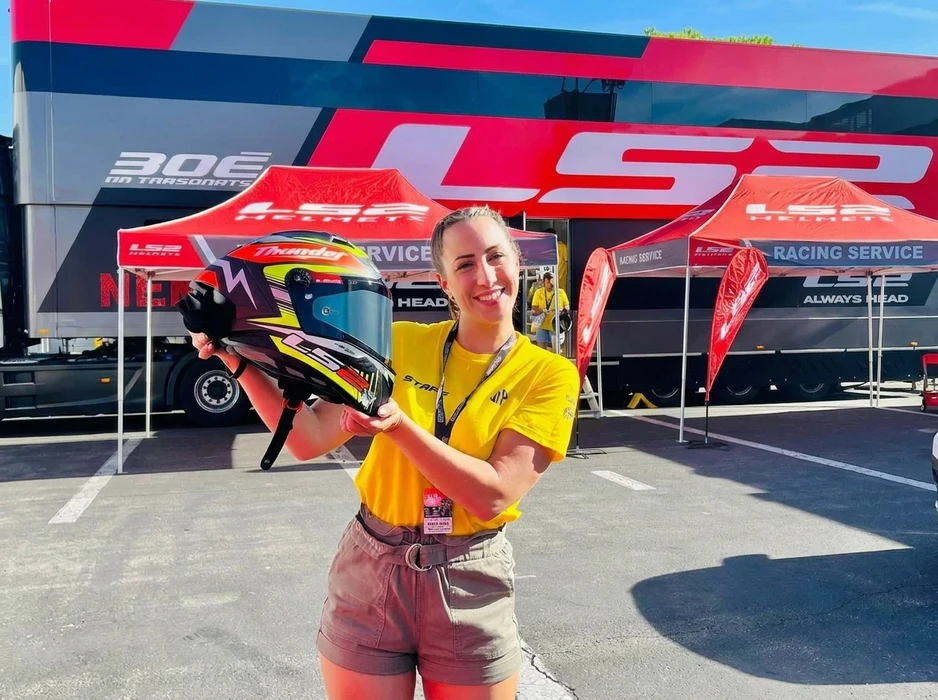 Femme souriante en t-shirt jaune tenant un casque moto LS2 Thunder, devant un stand de service racing LS2 Helmets
