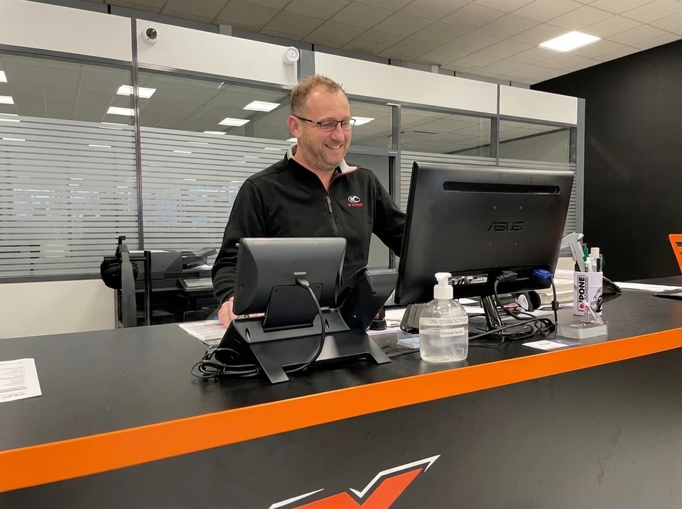 Homme souriant en polo noir debout derrière un comptoir de réception noir et orange, consultant un écran ASUS tout-en-un