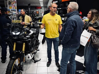 Photographie en intérieur du showroom/atelier d'Atout Motos. Trois à quatre membres de l'équipe en vêtements professionnels (polos ou chemises de travail aux couleurs de la marque) interagissent naturellement dans l'espace : un technicien en atelier présentant un élément de maintenance sur une moto sur béquille, un vendeur conseillant un client devant le comptoir, un collaborateur triant des équipements de protection. Ambiance lumineuse, accueillante et professionnelle, showcasing la cohésion de l'équipe et le contexte de métier : réparation, conseil client, équipement moto. Pas de logo ni texte visible.